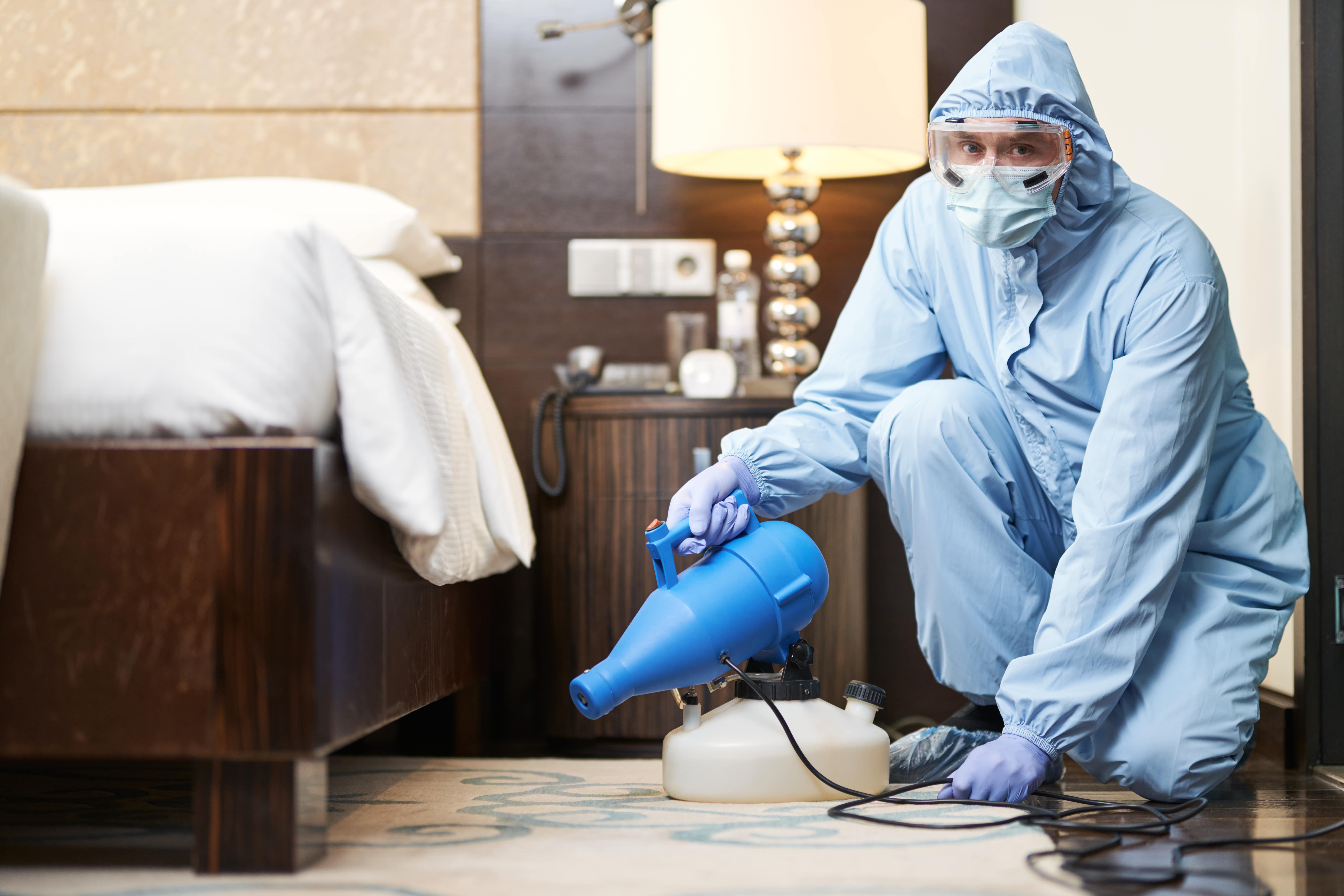 worker-in-biohazard-suit-and-mask-disinfecting-bed-min